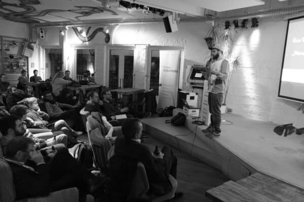 Ein Mann steht auf einer Bühne und spricht in ein Mikrofon. Vor der Bühne sitzen mehrere Menschen.