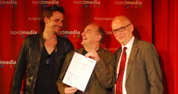 Drei Männer posieren für das Bild, einer von ihnen hält eine Urkunde in der Hand.