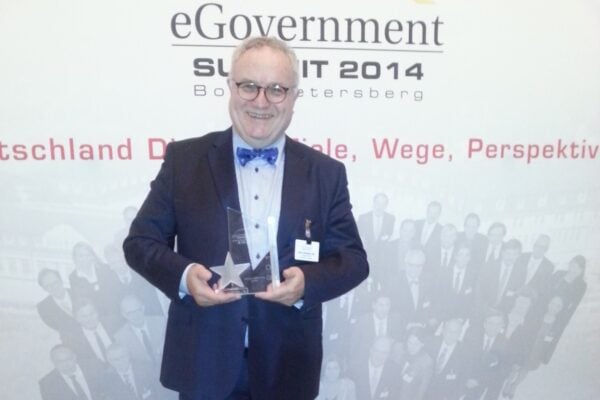 Ein Mann steht im Anzug mit einem Award aus Glas vor einer Fotowand und lächelt in die Kamera.