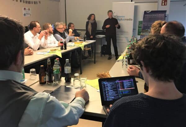 Mehrere Personen sitzen an Tischen in U-Form, vor diesem stehen zwei Personen die sprechen.