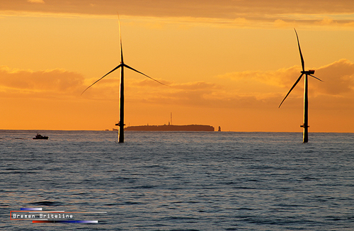 Zwei Windkrafträder stehen im Meer, der Himmel ist orange gefärbt.