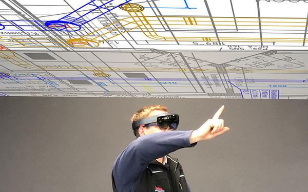 Ein Mann trägt eine VR-Brille und gestikuliert, darüber ein digitaler Bauplan
