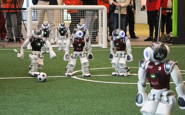 Roboter spielen auf einem künstlichen Fußballfeld; im Hintergrund ist ein Tor zu sehen.