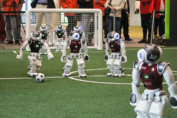 Roboter spielen auf einem künstlichen Fußballfeld; im Hintergrund ist ein Tor zu sehen.