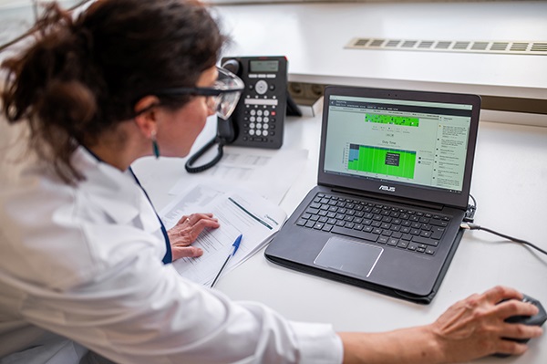 Eine Frau in weißem Laborkittel und mit Schutzbrille ist über einen Laptop gebeugt und guckt auf den Bildschirm.