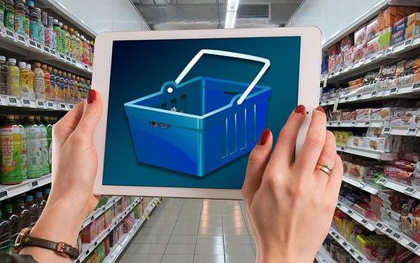 Hände halten in einem Supermarkt-Gang ein iPad auf dem ein digitaler Einkaufskorb zu sehen ist.