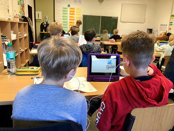 Blick in den Klassenraum von der Rückseite aus. Im Fokus zwei Jungen die gemeinsam auf ein Tablet gucken.
