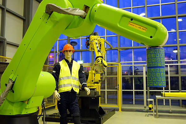 Ein grüner Roboterarm steht im Vordergrund, dahinter eine Schaufensterpuppe mit gelber Warnweste.
