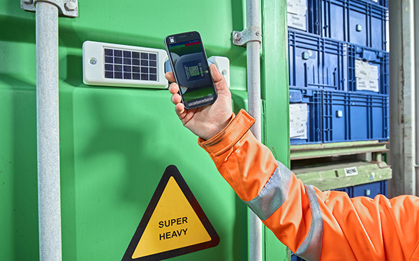 Arm in orangefaarbener Schutzkleidung hält ein Smartphone vor einen grünen Container.