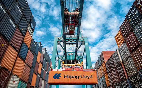 Containerarm mit einem orangefarbenem Hapag-Lloyd Container im Griff. Links und rechts sind ebenfalls zahlreiche Container aufgestapelt.