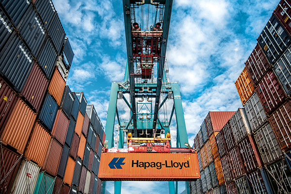 Containerarm mit einem orangefarbenem Hapag-Lloyd Container im Griff. Links und rechts sind ebenfalls zahlreiche Container aufgestapelt.