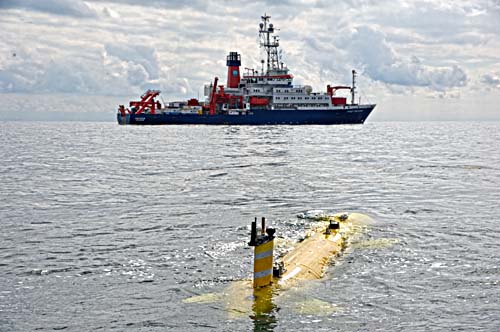 Im Wasser fährt ein autonomer, gelber Tauchroboter, im Hintergrund sieht man ein großes Schiff.