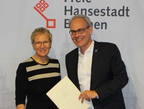 Ein Mann und eine Frau posieren für das Bild, im Hintergrund sieht man das Logo der Stadt Bremen.