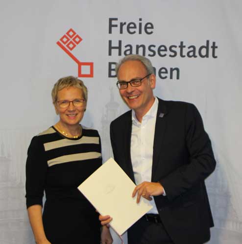 Ein Mann und eine Frau posieren für das Bild, im Hintergrund sieht man das Logo der Stadt Bremen.