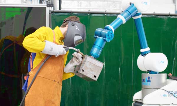Ein Roboterarm hät einen Gegenstand, an dem eine Person in gelber Schutzkleidung arbeitet.