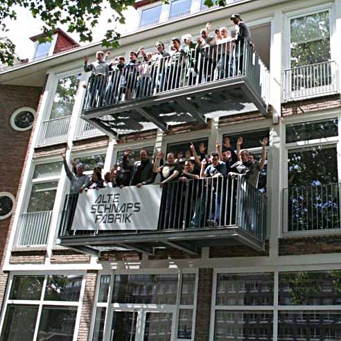 Foto das eine Hauswand mit zwei Balkonen zeigt auf denen viele Menschen stehen und in die Kamera winken.