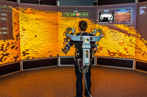 Eine Person ist verkabelt und steht vor mehreren Bildschirmen, auf denen eine Marsmission simuliert wird.