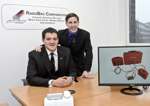 Zwei Männer posieren neben einem Bildschirm für das Bild, im Hintergund ist ein Logo zu sehen.