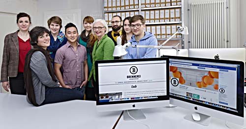 Eine Gruppe aus Männern und Frauen steht hinter zwei Computerbildschirmen und schaut in die Kamera.