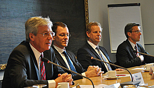 Vier Männern sitzen in einer Pressekonferenz an einer Tischreihe mit Mikrofonen, einer spricht.