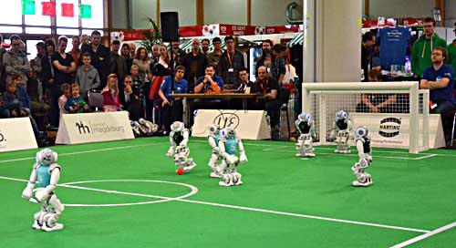 Auf einem Kunstrasefeld spielen mehrere Roboter mit einem Fußball, darum herum stehen viele Zuschauer.