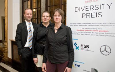 Drei Personen posieren vor einem Roll-Up vom Bremer Diversity Preis.