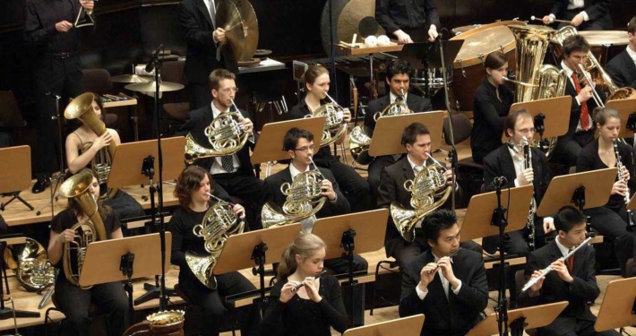 Junge Menschen sitzen in einem Orchester zusammen und spielen verschiedene Blasinstrumente.