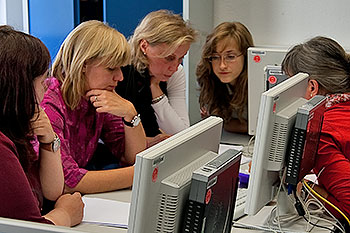 Drei Frauen gucken sich gemeinsam etwas vor sich auf dem Tisch an; vor ihnen im Bild stehen Computer.
