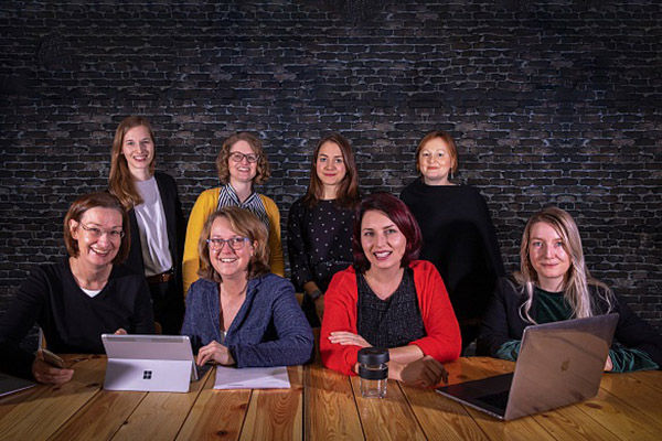 Acht Frauen schauen in die Kamera und haben Laptops vor sich stehen