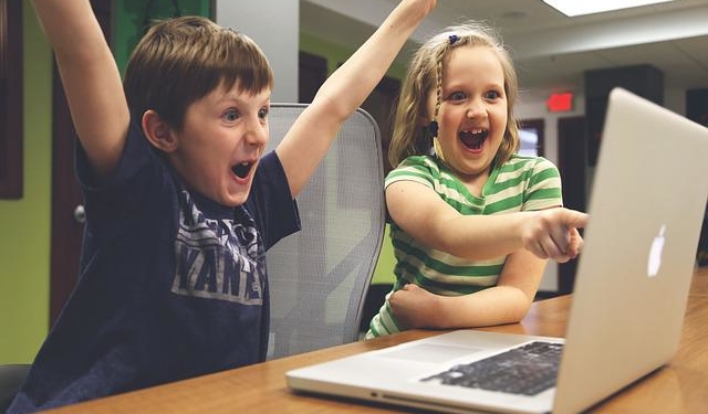 Ein Junge und ein Mädchen sitzen mit begeistertaufgerissenen Augen vor einem Laptop. Der Junge reißt die Arme in die Luft, das Mädchen zeigt auf den aufgeklappten Bildschirm.