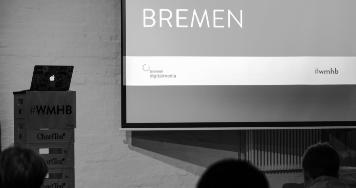 Schwar-weiß Bild eines Bühne mit Beamer-Projektion, darauf der Schriftzug: Webmontag Bremen