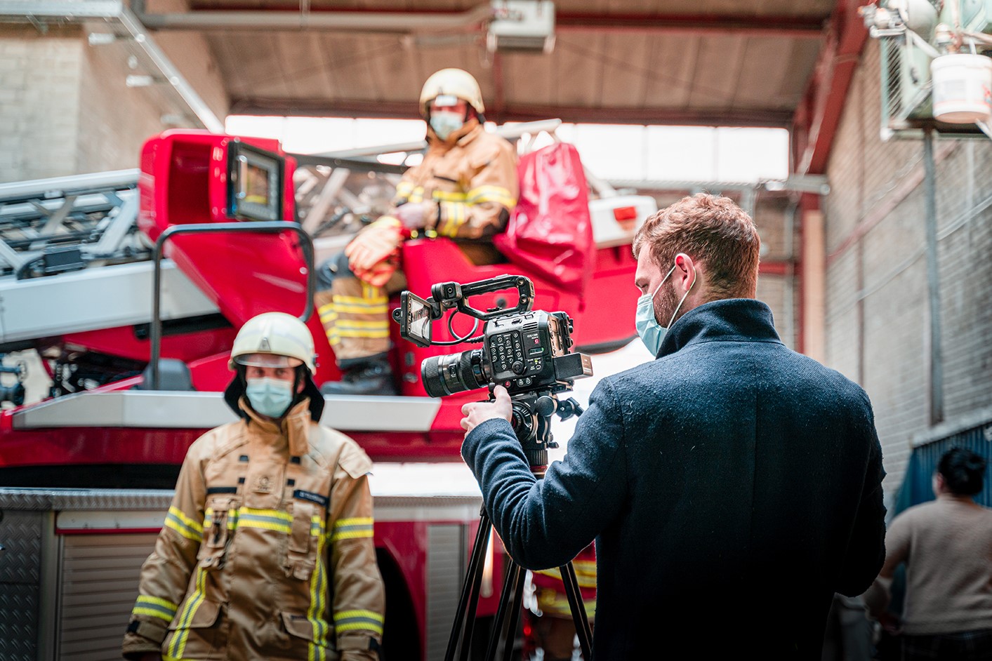 Zwei Feuerwehrleute in Monturwarten auf Anweisungen eines Filmemachers im Vordergrund der hinter seiner Kamera steht.