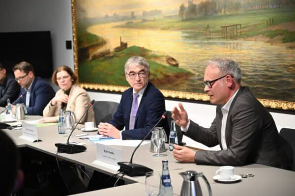 Vier Menschen sitzen mit Mikrofonen an einer Tischreihe; der Mann vorne im Bild spricht.