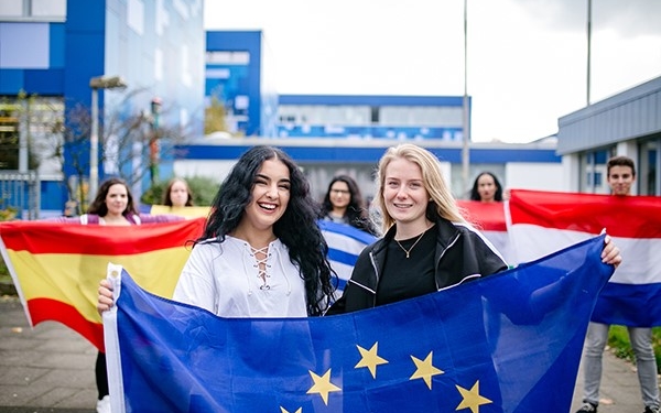 Junge Menschen halten Fahnen; immer zwei ein. Im Vordergund zwei junge Frauen, die eine Europaflagge halten.