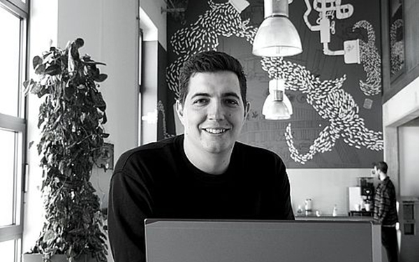 schwarz-weiß-Foto eines Manes der hinter einem Laptop sitzt und in die Kamera lächelt.