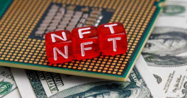 Drei rote Würfel bilden die Buchstabenfolge NFT. Sie liegen auf einer Leiterplatine und auf US-Geldscheinen.