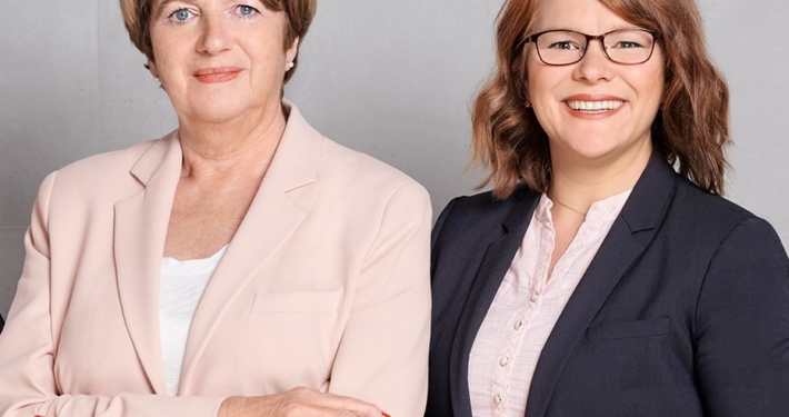 Zwei Frauen vor grauem Hintergrund lächeln in die Kamera.