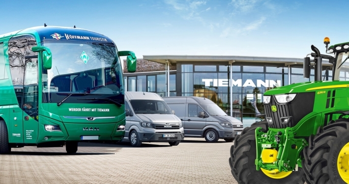 Ein Furpark an großen Fahrzeugen: Ein Bus, ein Trakor und zwei Sprinter, im Hintergrund ein Gebäude mit Glasfront, darauf der Schriftzug Tiemann.