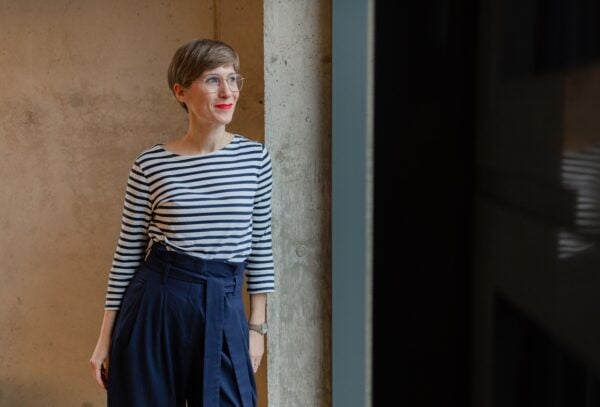 Foto einer Frau die innen an einer Mauer lehnt und nach rechts aus dem Bild und einem Fenster guckt.