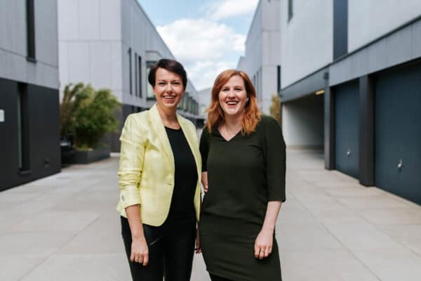 Zwei Frauen stehen in einer Häuserschlucht und lächeln in die Kamera.