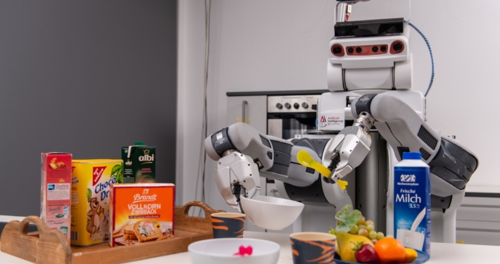 Ein Roboter steht mit Plastiklöffel und Schale in den "Händen" an einem Tresen Auf dem Tresen sind verschiedene Lebensmittel.
