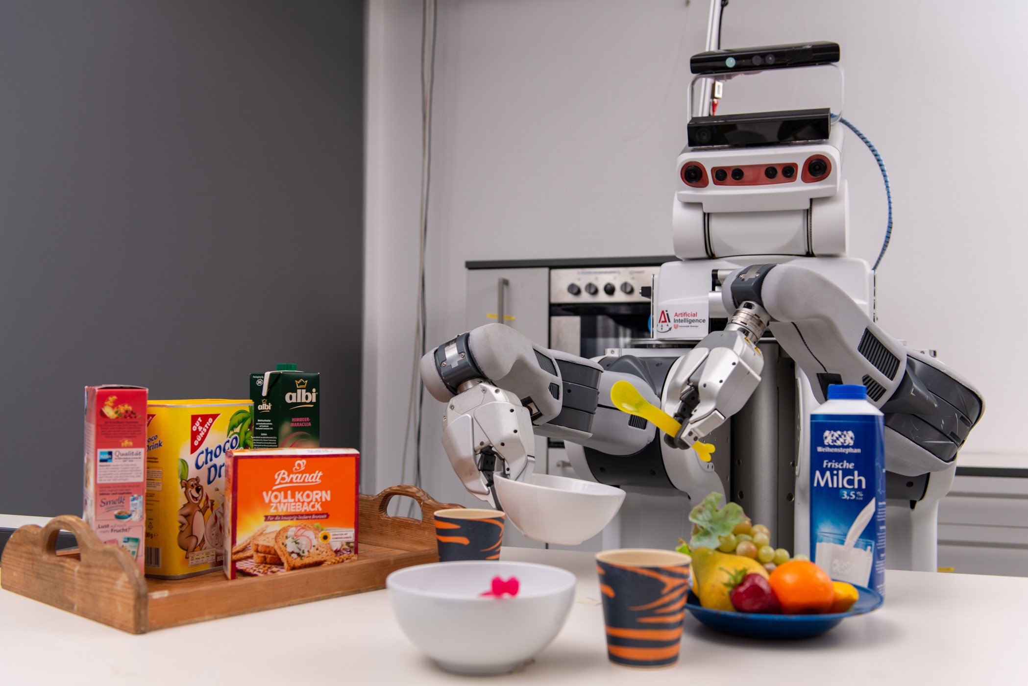 Ein Roboter steht mit Plastiklöffel und Schale in den "Händen" an einem Tresen Auf dem Tresen sind verschiedene Lebensmittel.