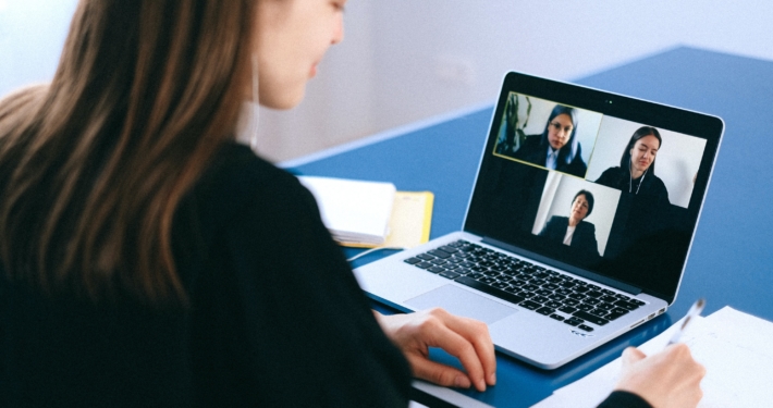 Eine Frau sitzt seitlich mit dem Rücken zur Kamera und schaut auf einen Laptop, der drei Personen zeigt, die gerade in einem Meeting mit ihr sind.