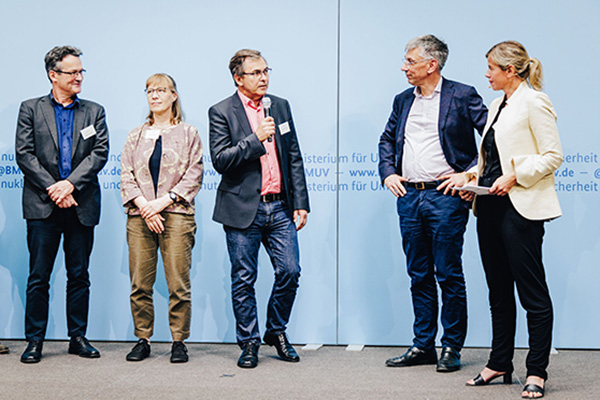 Fünf Menschen stehen auf einer Bühne vor einer Werbewand. Ein Moderator in der Mitte stellt gerade einer anderen Person einer Frage.