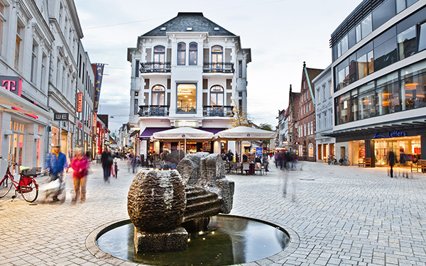 Blick auf einen belebten Platz im Stadtinneren mit Häusner, Geschäften und einem Skulptur-Brunnen innder Mitte.