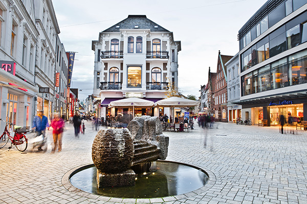 Blick auf einen belebten Platz im Stadtinneren mit Häusner, Geschäften und einem Skulptur-Brunnen innder Mitte.