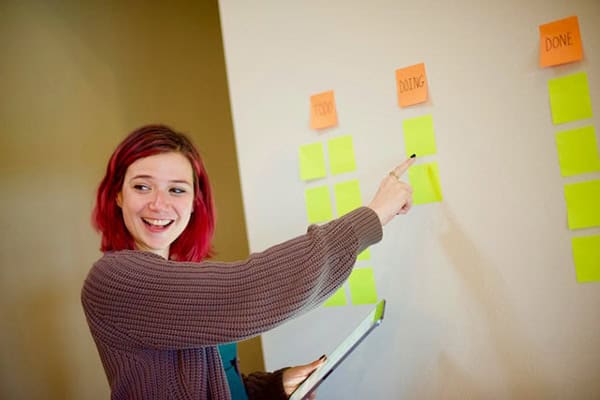 Einen Frau steht neben einem Whiteboard und zeigt den Blick vom Board abgewandt hin zu einem nicht im Foto sichtbaren Punkt auf verschiedene PostIts die am Board kleben.