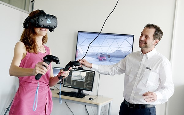 Eine Frau mit VR Brille und Steuerungselementen in der Hand steht neben einem Mann, derdie Frau beobachtet. Im Hintergrund sind Bildschirme zu sehen.