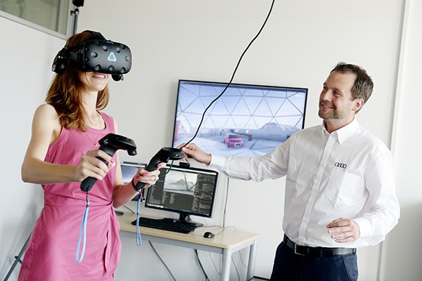 Eine Frau mit VR Brille und Steuerungselementen in der Hand steht neben einem Mann, derdie Frau beobachtet. Im Hintergrund sind Bildschirme zu sehen.