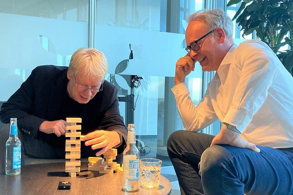 Zwei Männer sitzen an einem runden Couchtisch und gucken auf einen Jenga Turm aus gestapelten kleinen Holzklötzen. Einer der beiden zieht einen Klotz aus dem Turm.